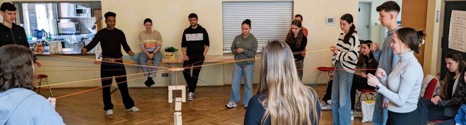 mehrere Jugendliche spiele "Tower of Power". Sie halten Schnüre in den Händen und müssen gemeinsam einen Haken mit Bausteinen daran balancieren